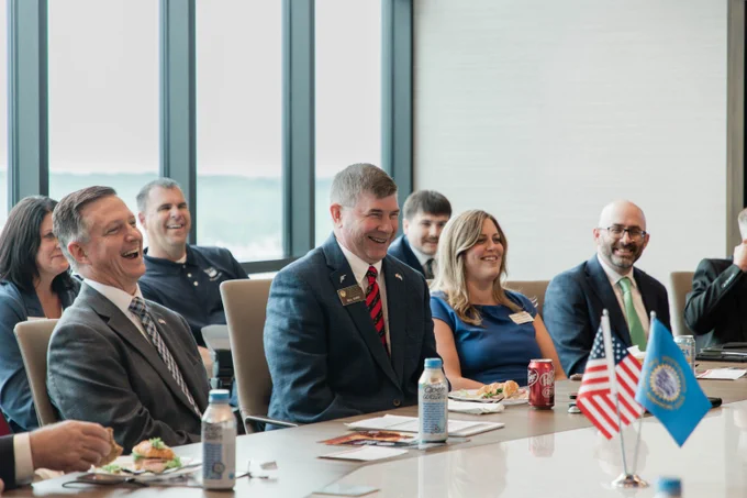 A group of people sitting at a conference table, smiling and engaging in conversation. Two small flags, including the United States flag, are placed on the table.