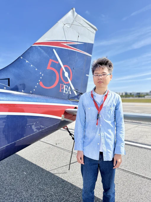 A person stands beside the tail of a small aircraft on a sunny day. The aircraft tail features a "50 PEA" logo with red, white, and blue accents. The person is wearing glasses, a light blue shirt, and a red lanyard. The background shows an airfield with a clear blue sky.