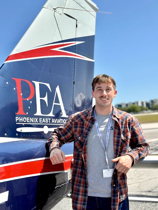 A person standing next to a small aircraft with "Phoenix East Aviation" and the initials "PEA" on the tail. The scene is outdoors with a clear blue sky.