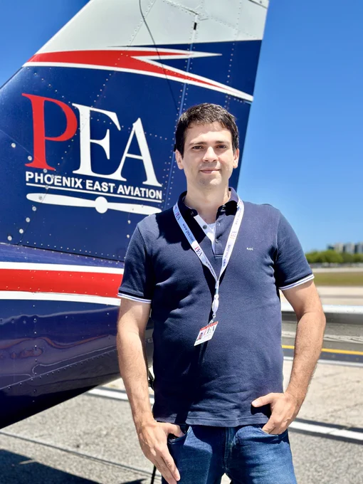 A person is standing in front of an aircraft with the Phoenix East Aviation logo on the tail. The background includes a clear blue sky and part of an airport runway.