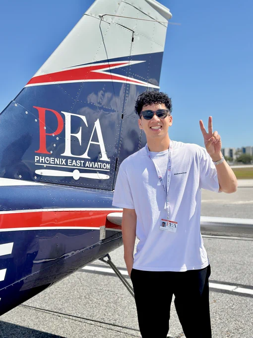 Person standing in front of a Phoenix East Aviation airplane, smiling and showing a peace sign, with a clear blue sky in the background.