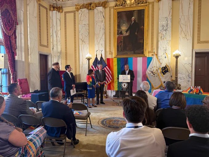 photo of crowd in state room watching secretary amore speak at podium with pride flags in background