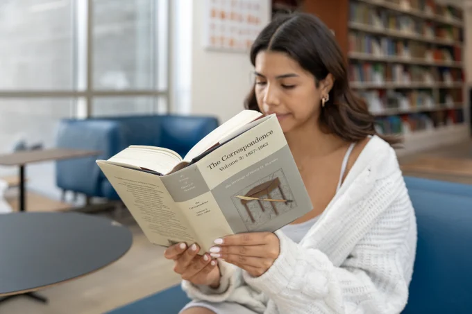 A UCSB student reads "Correspondence Volume 3." The student is seated on a blue lounge chair in the Sara Miller McCune Arts Library. A bookshelf is visible in the background.