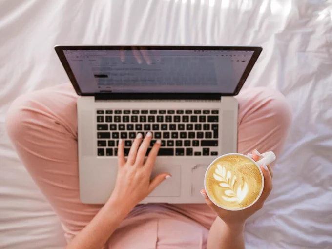 Person with pink pants with a laptop on their lap typing with their left hand, holding a latte in their right. Latte has a leaf design.