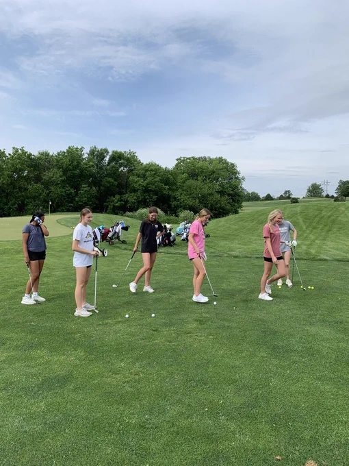 Five people stand on a golf course. They hold golf clubs. Some wear white shoes. Trees and a cloudy sky are in the background.