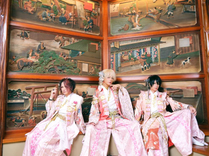 Seth, Kouki and Tsukasa of Sakura Zensen dressed in traditional kimono with hakama, with pink and red hues and floral patterns. They are seated side by side with Japanese (wooden) artworks in the background. © Sakura Zensen