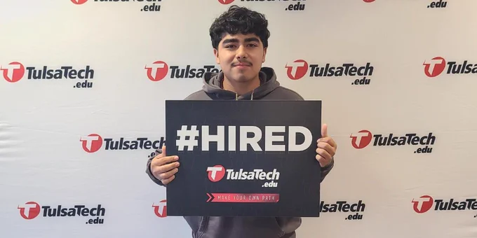 Person standing in front of a Tulsa Tech backdrop holding a #HIRED sign, celebrating job placement success.