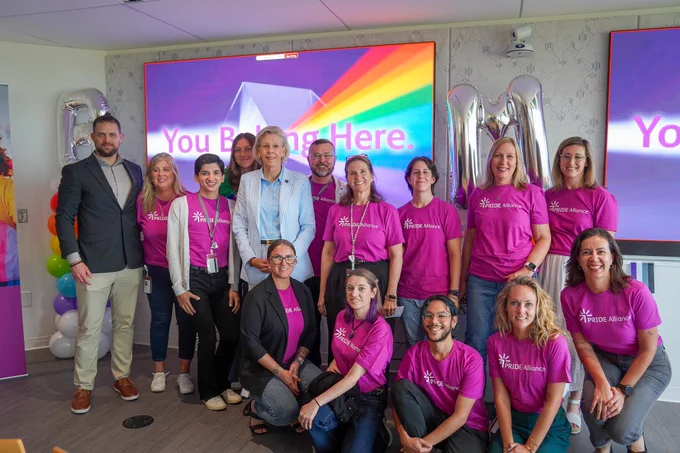 Mayor Jane Castor stands together with employees from Bristol Myesr Squibb. in a brightly lit room. Several individuals wear matching purple shirts bearing a logo with the words "Pride Alliance." Behind them is a screen displaying a rainbow design and the words "You Belong Here.