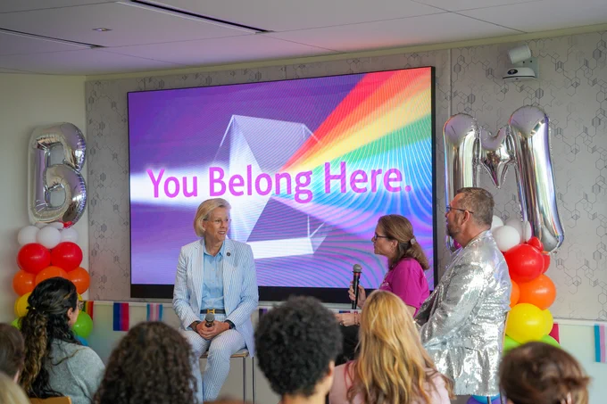 A panel discussion is taking place in a room with a diverse audience. A large screen displays the message "You Belong Here" with colorful graphics. Two inflatable balloons with the letters "B" and "M" are visible. Three individuals are seated in front of the screen, with one holding a microphone.