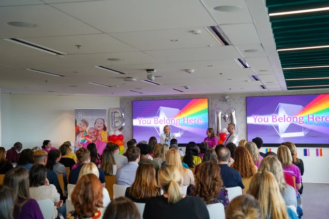 A group of people seated in a conference room faces a panel of speakers. Two large screens at the front display the message "You Belong Here" with rainbow graphics. Balloons and international flags decorate the space, creating a welcoming atmosphere.