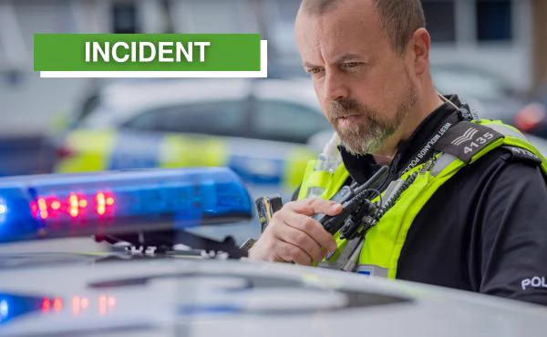 A West Midlands police officer with a radio, next to a police car with flashing blue lights.