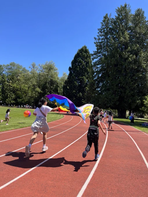 Two children running on a red track under a clear blue sky, one holding a pride flag behind them.