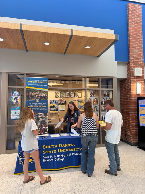 A person standing behind a table with a South Dakota State University Honors College banner, talking to a new student and her parents. The setting is an indoor hallway with posters on the walls.