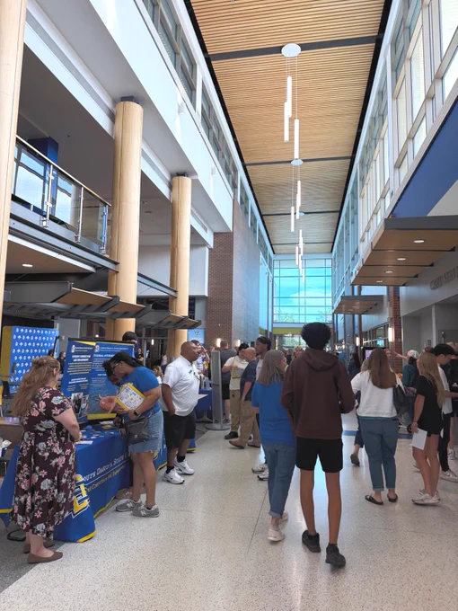New Student Orientation in the SDState Student Union features attendees visiting information booths. South Dakota State University is prominently displayed on signs at one booth, while people walk, chat, and collect brochures. Tall ceilings and large windows illuminate the scene.