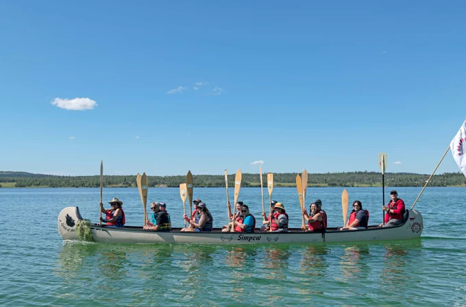 Group of nine people paddling a long canoe on calm lake water under a clear blue sky.
----
Un groupe de neuf personnes pagayant dans un long canoë sur un lac aux eaux calmes, sous un ciel bleu dégagé.
