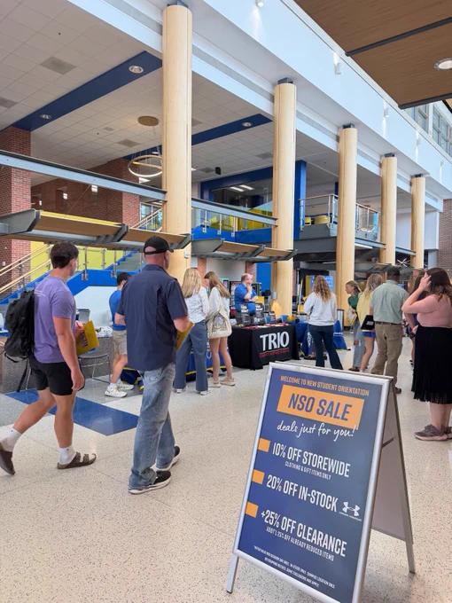 A group of people stand in a university building lobby. There's a table with a "TRIO" sign, and others walk or chat. A poster in the foreground advertises a sale at Jackrabbit Central, the school bookstore, highlighting discounts including 10% off storewide, 20% off in-stock items, and 25% off clearance, featuring Under Armour. The area is spacious with large windows and pillars.