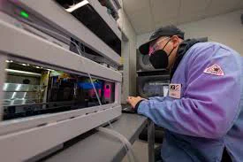 An individual wearing a mask and glasses works on a complex machine. The person is focused and surrounded by various electronic equipment.