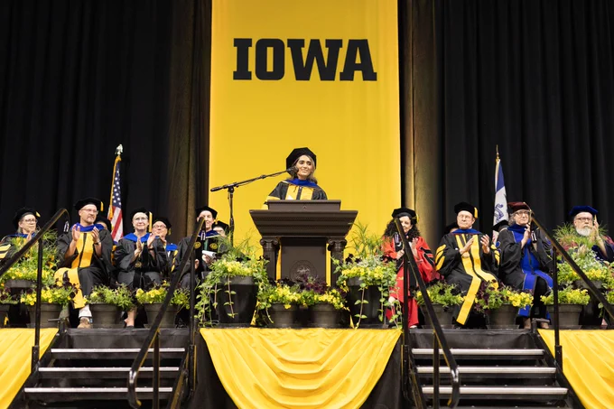 University of Iowa graduation ceremony with faculty in academic regalia seated on stage behind a speaker at the podium.