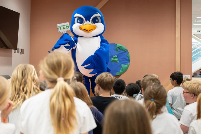 A large blue mascot entertains a group of kids in a lively setting, holding a sign that says 'YES.' The children appear attentive and excited.