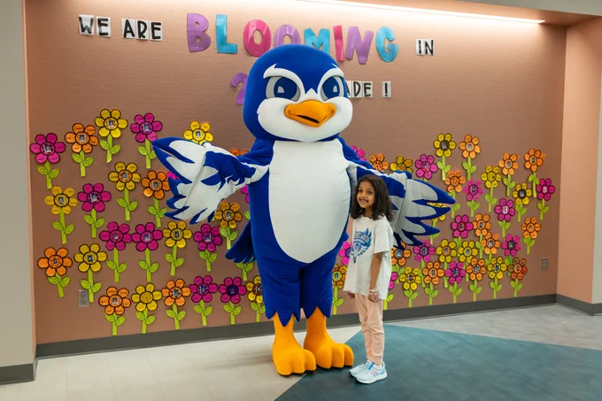 A young girl smiles while posing with a large, blue bird mascot in front of a colorful flower decor with a cheerful atmosphere.