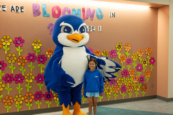 A cheerful school scene featuring a blue mascot standing beside a smiling student. They are in front of a colorful wall with paper flowers and the words 'We are Blooming in' in large playful letters. The mood is joyful and celebratory.