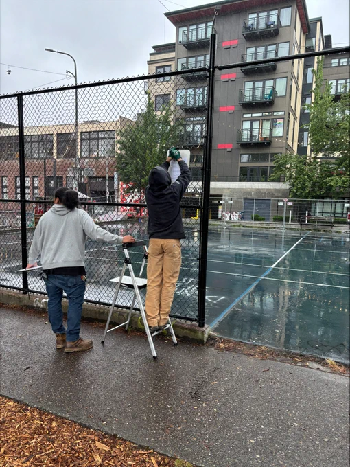 Two people working on a chain-link fence beside a wet outdoor basketball court in an urban area with apartment buildings in the background.