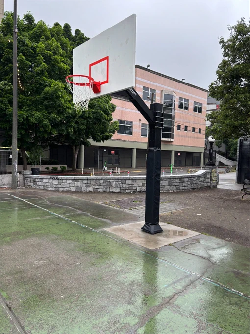 Outdoor basketball hoop with a red rim and white backboard on a wet concrete court near a school building.