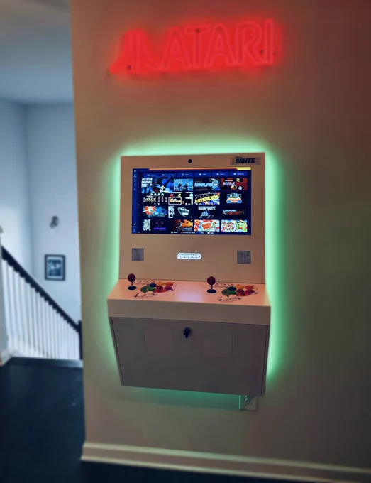 Wall-mounted Polycade Sente machine with illuminated screen displaying classic games and colorful joystick controls under a glowing red Atari sign.