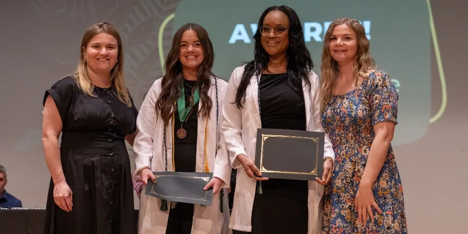 Four people stand on stage during an award ceremony, two wearing white coats holding plaques and medals, with a presentation screen behind them.