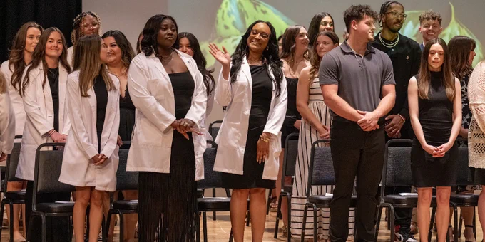 Group of students standing on stage during the health professions ceremony, some wearing white coats and formal attire.