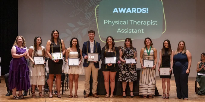 Group of 10 individuals standing on stage holding certificates under a sign reading "AWARDS! Physical Therapist Assistant."