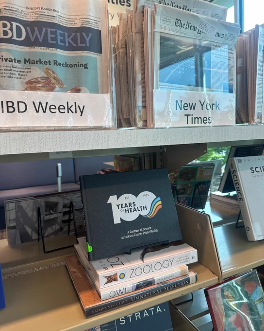 A shelf displays various newspapers and books. Newspapers include "The New York Times." Books on the shelf feature titles like "100 Years of Health" by Jackson County Public Health, among others.