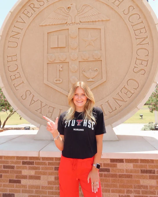 Female nursing student wears red scrub pants and a black TTUHSC t-shirt and stands outside in front of the university seal.