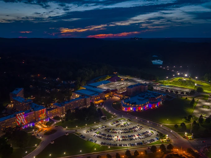 Aerial view of the brightly lit French Lick Resort at dusk, surrounded by a large parking area with numerous cars. The sky is dark blue with a hint of orange near the horizon, and the landscape includes visible trees and open fields.