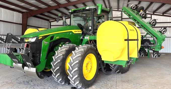 A green John Deere tractor with yellow wheels is parked inside a metal-roofed warehouse. It is attached to a large piece of farming equipment with multiple wheels and a yellow component labeled "Nurse.