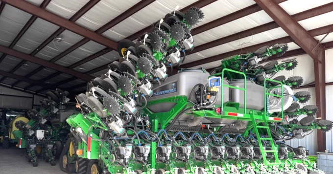 A large John Deere farming machine is stored in a spacious metal shed. It features multiple rows of disc blades and green and black components, with the John Deere logo visible. The shed roof is supported by metal beams, and there are other farming equipment partially visible in the background.