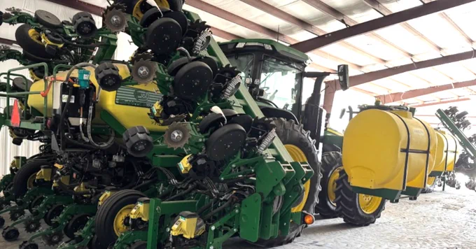 A large John Deere tractor is parked inside a building. It is equipped with farming attachments, including multiple rows of wheels and a large yellow tank on one side. The roof structure is visible, along with a concrete floor underneath.