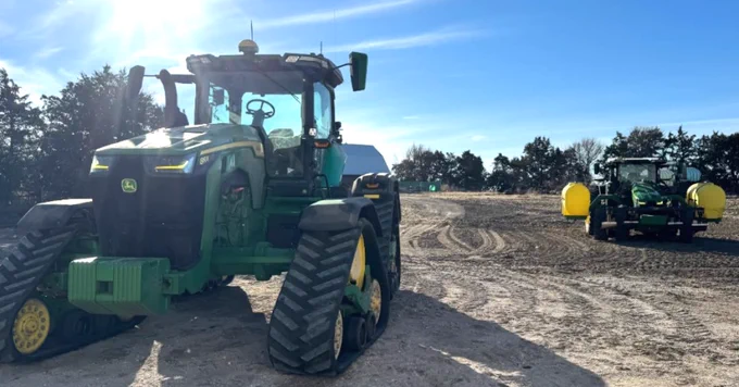 A John Deere tractor with large tires is positioned on a dirt road under a bright sun. Another similar tractor is visible in the background. Surrounding the scene are trees beneath a clear blue sky.
