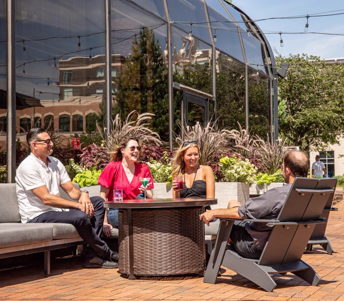 Four people sit around an outdoor table, laughing and enjoying colorful drinks. They're in a sunny, urban setting with plants and a glass-domed building nearby.