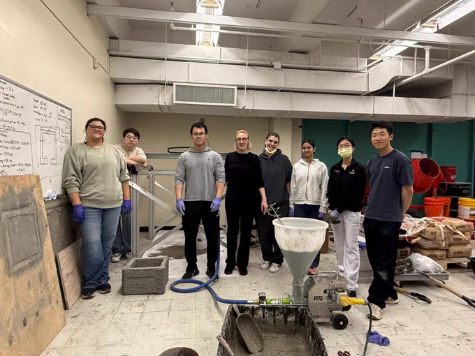 A group of eight people standing in a workshop, surrounded by industrial equipment, bags of materials, and construction tools. Some are wearing gloves, and others have safety glasses. A whiteboard with sketches and notes is on the wall to the left. The environment is busy and industrial.