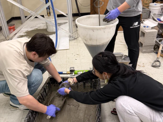 Three individuals are working with wet concrete in a laboratory setting. Two are kneeling near a large black tray, smoothing the concrete with trowels, while the third is standing, holding a pipe connected to a large funnel. Various tools and concrete blocks are visible in the background.