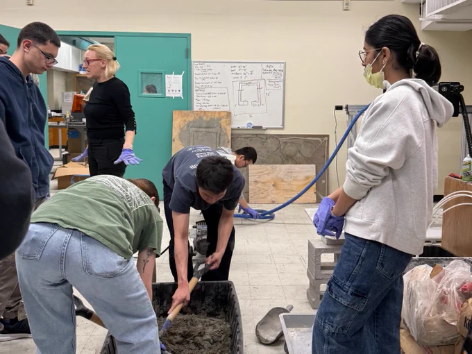 A group of individuals is engaged in a workshop or educational setting. One person is mixing a substance in a container with a tool, while others observe or assist. A whiteboard with drawings and notes can be seen in the background, along with various pieces of equipment and materials.