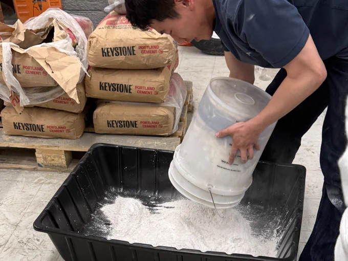 A person is pouring a white powdery substance from a clear plastic container into a large black bin. In the background, there are several bags labeled "Keystone Portland Cement" stacked on wooden pallets.