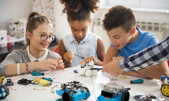 Several youth are gathered around a table, actively engaging in constructing small robotic devices together. The table is scattered with various electronic components and tools. The environment appears lively and educational.