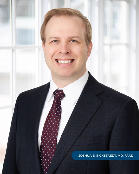 A professional portrait of Joshua B. Eickstaedt, MD, FAAD, wearing a dark suit and tie and smiling in front of a paned glass window