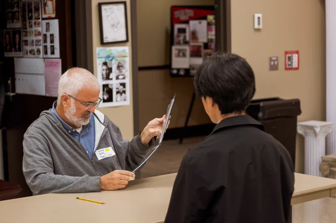 Older man with gray hair and glasses reviewing a document across a desk from a younger person in a black jacket.