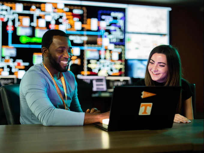 Two people sitting at a table, using a laptop with a Tennessee Volunteers logo. There are large screens with complex diagrams in the background. They appear engaged and focused.