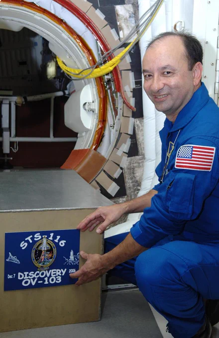 Mark Polansky, wearing a blue flight suit, smiles as he squats while placing a mission placard at the hatch opening in the White Room of Launch Pad 39B. The placard reads: "STS-116 Discovery OV-103."
