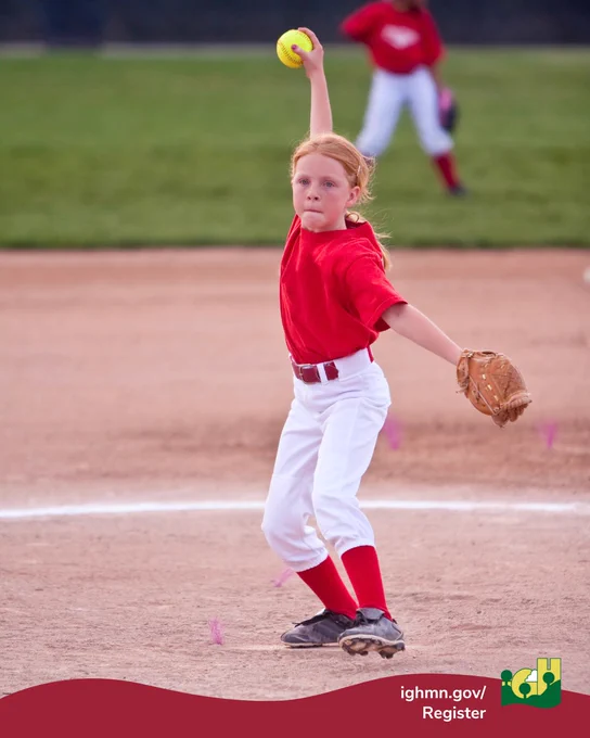 Young softball player in red jersey winding up to pitch on a dirt field with a teammate in the background.
