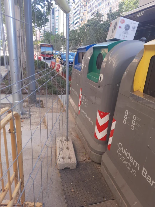 A la derecha una valla de obra en diagonal. A la derecha contenedor azul, verde y amarillo.
La valla cierra el paso al contenedor verde y al amarillo.